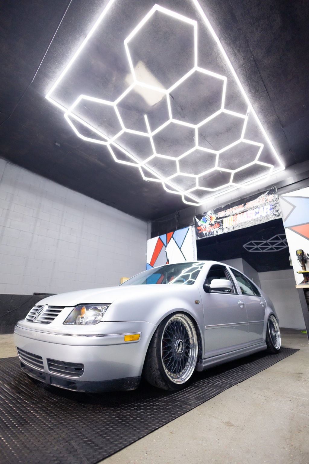 Silver Volkswagen Jetta with mesh wheels inside the shop under hexagonal lighting